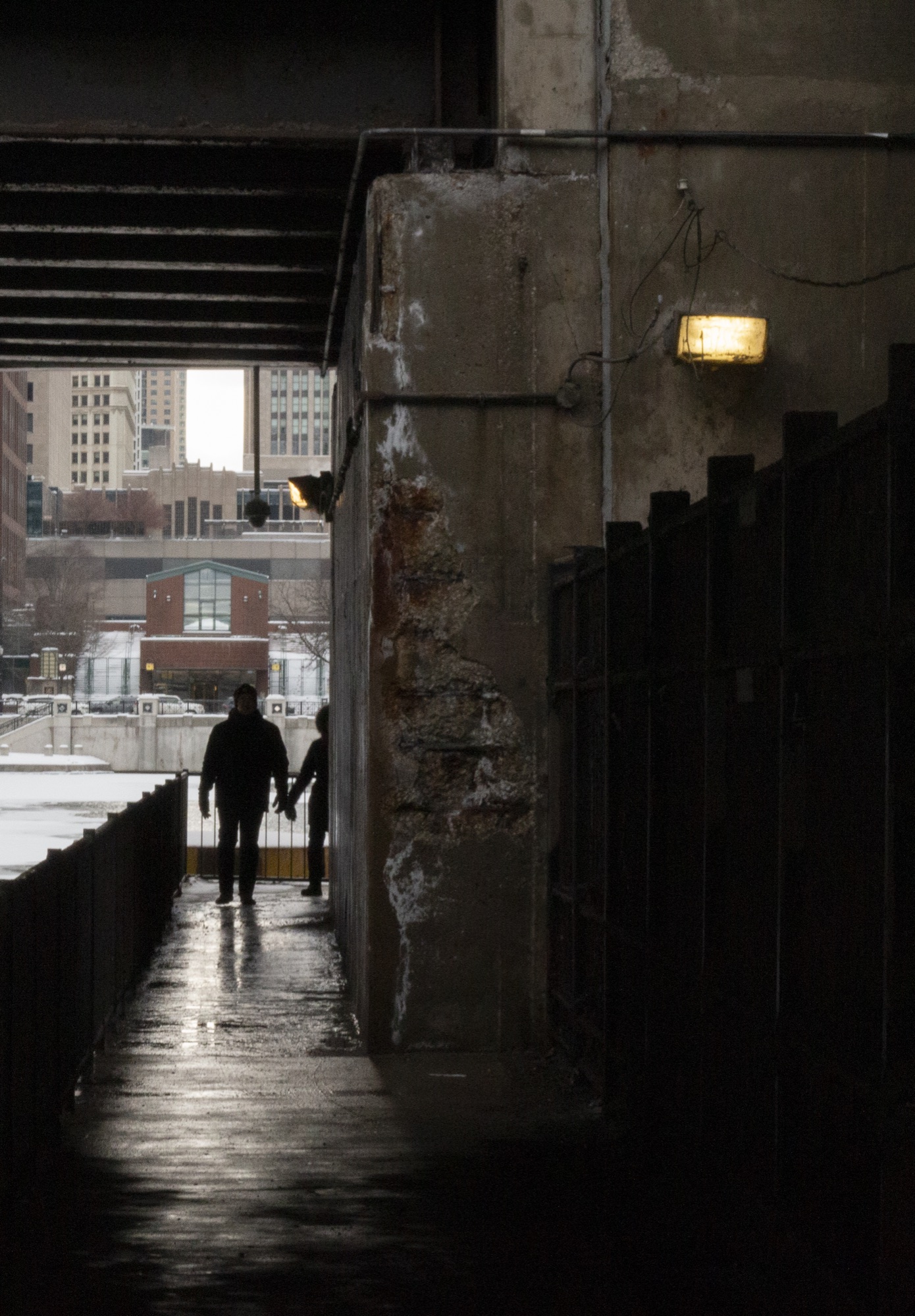 Two figures holding hands walking out of a dark riverside underpass toward snow and daylight, a bulb lit on the concrete pier above them