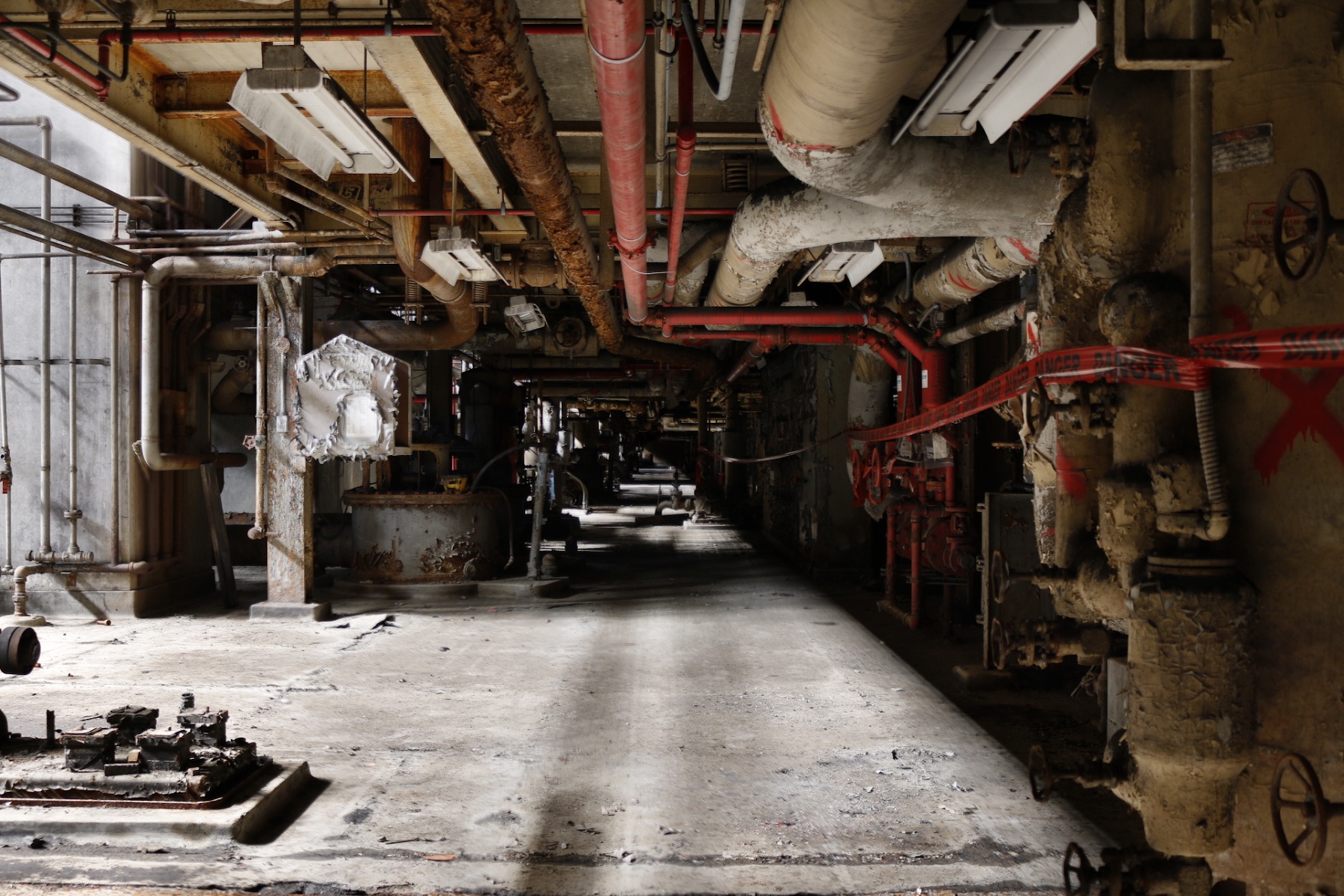 A basement corridor lined with lagged pipework and red-painted runs, daylight falling across the dusty concrete floor