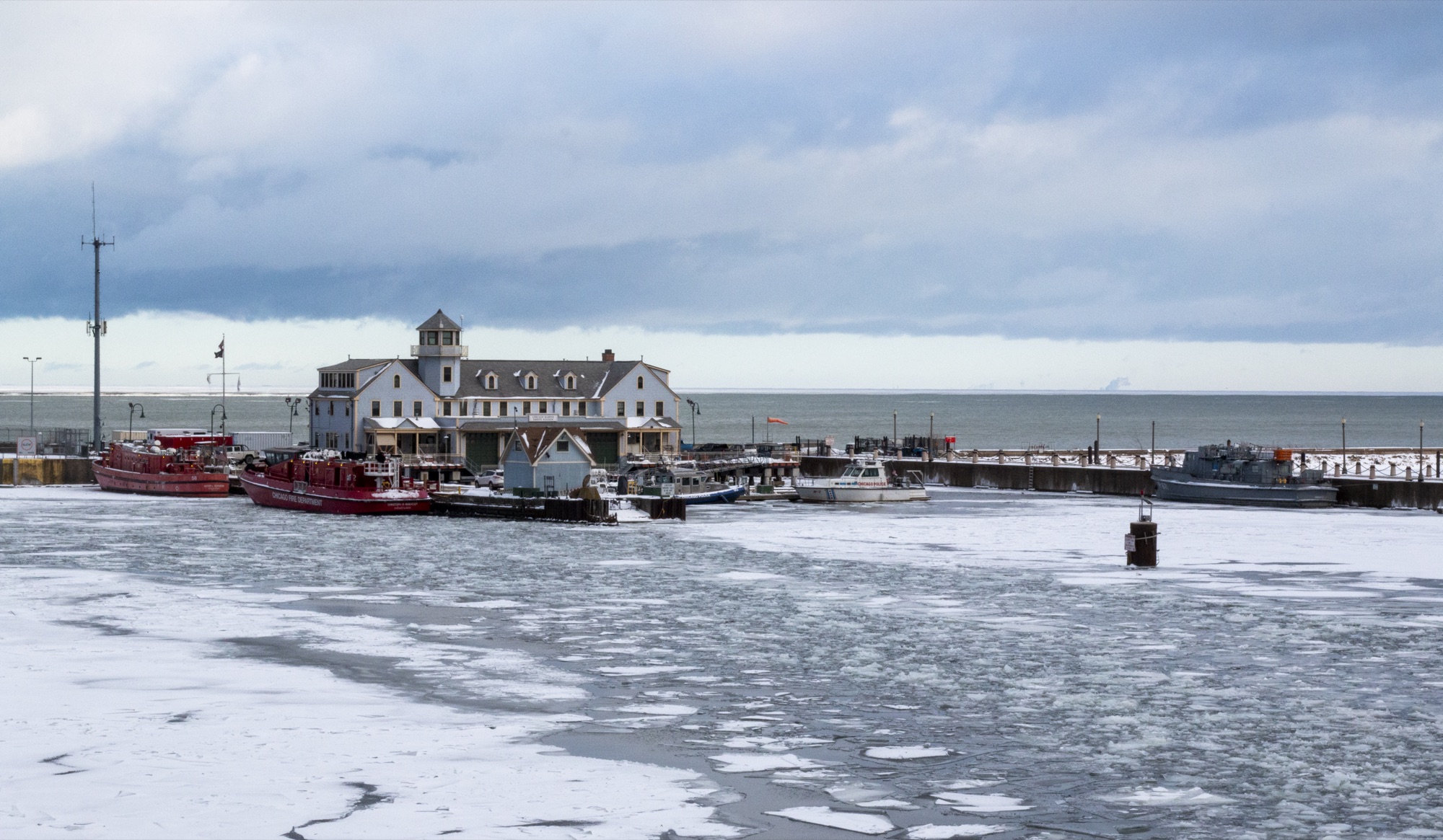 Ice covering a harbor in front of a white lakefront station building, fireboats and tugs moored alongside