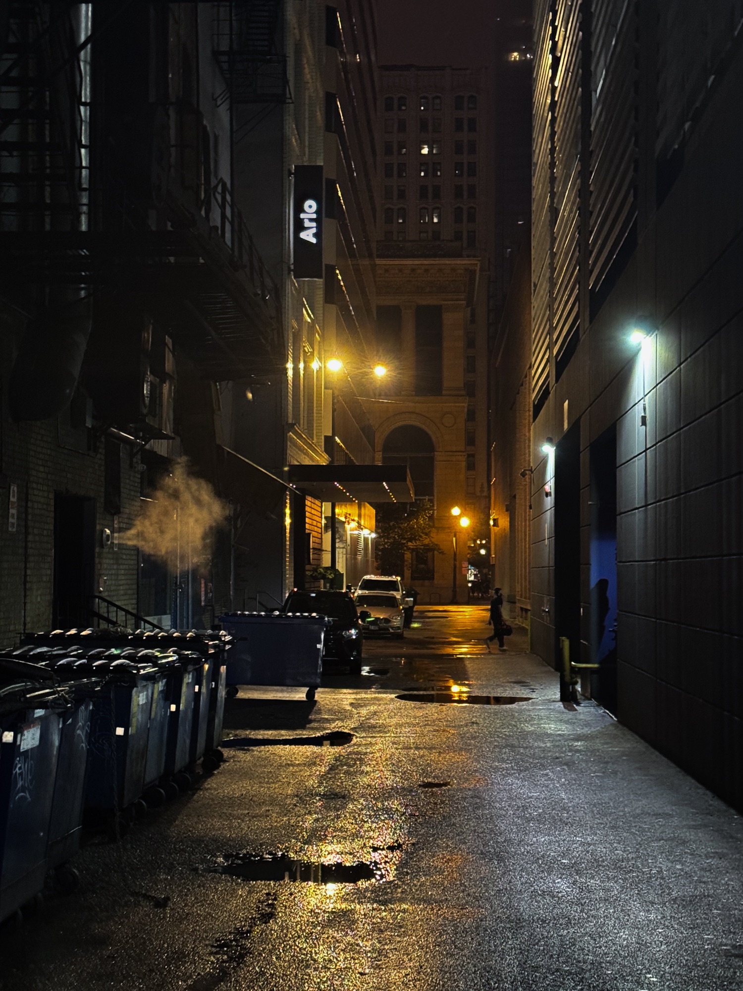 A wet downtown alley at night, steam rising past dumpsters and warm streetlight reflected off the pavement, a pedestrian walking out of view