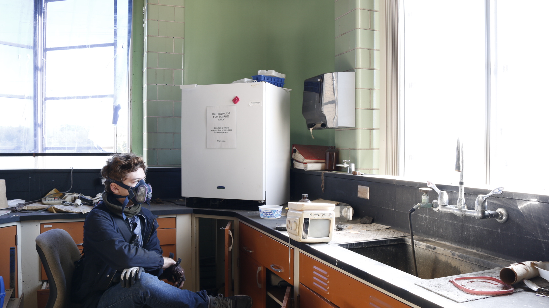 A person in a respirator sitting in an old chair in an abandoned laboratory kitchen, cabinets and a deep sink alongside