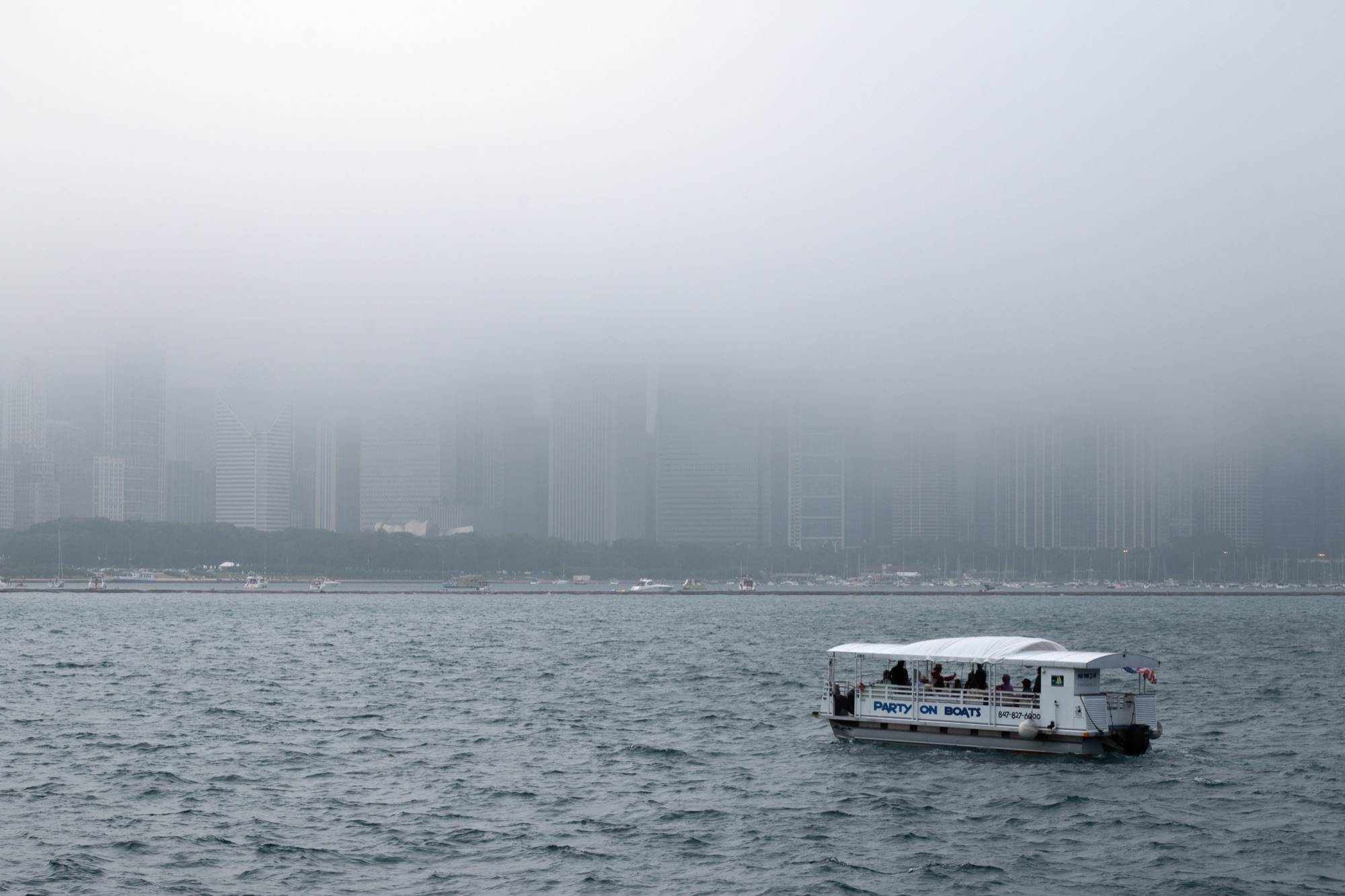 A small party boat on a slate-gray lake heading towards the Chicago skyline, which dissolves into pale fog