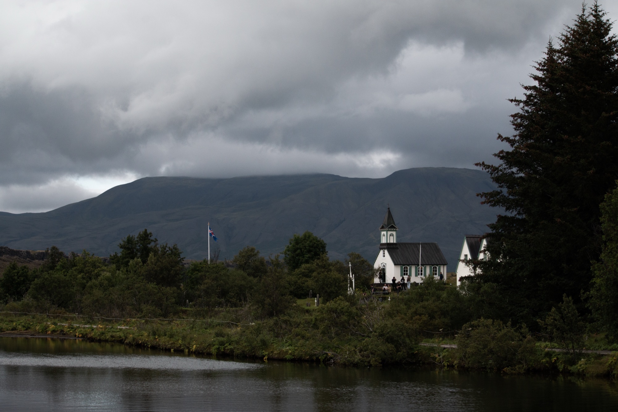 A small white church with a dark steeple beside a still river, heavy cloud massed over a flat-topped mountain