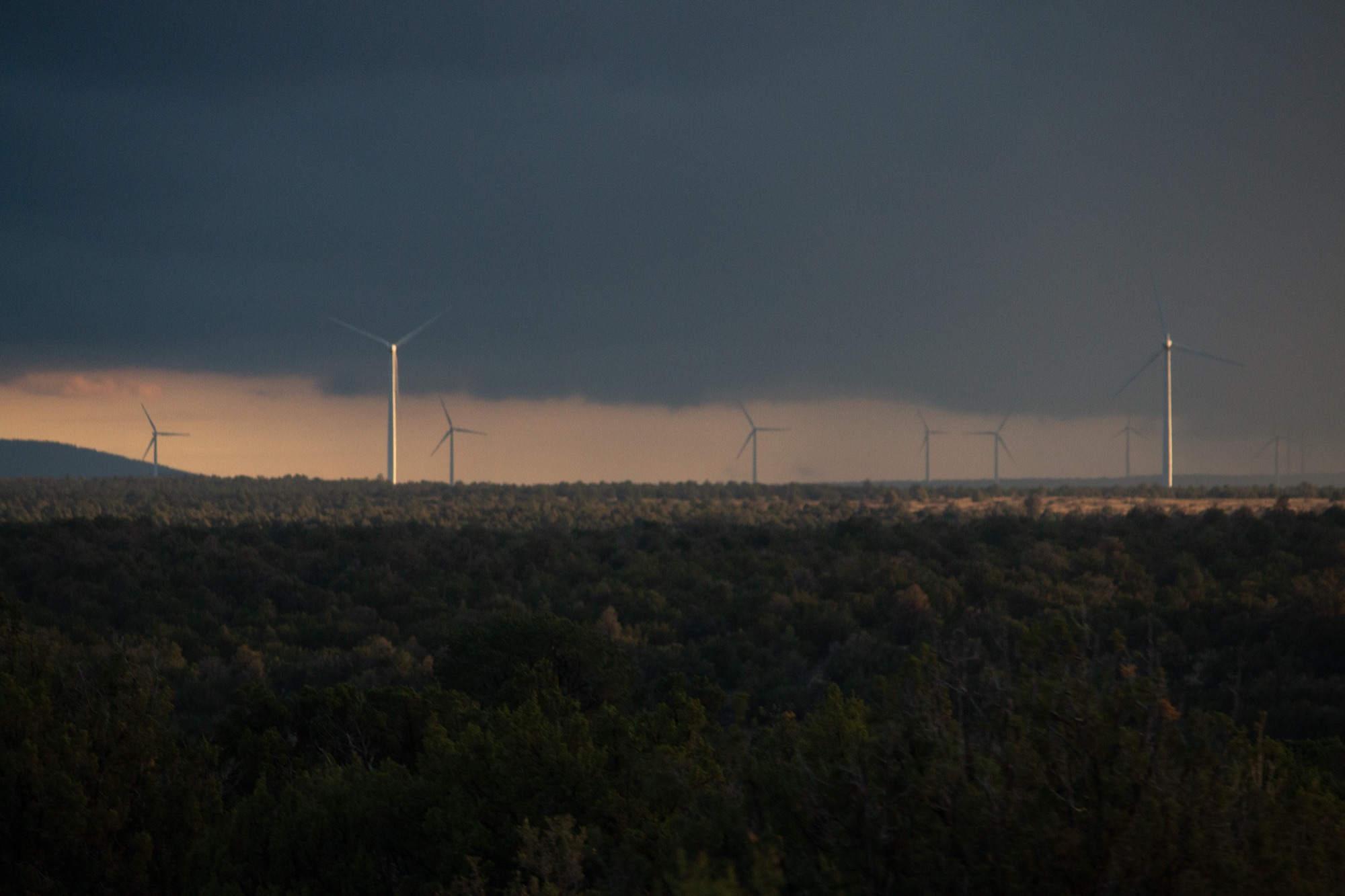 Wind turbines strung along a dark forested ridge beneath a storm front, a narrow band of dull orange light at the horizon