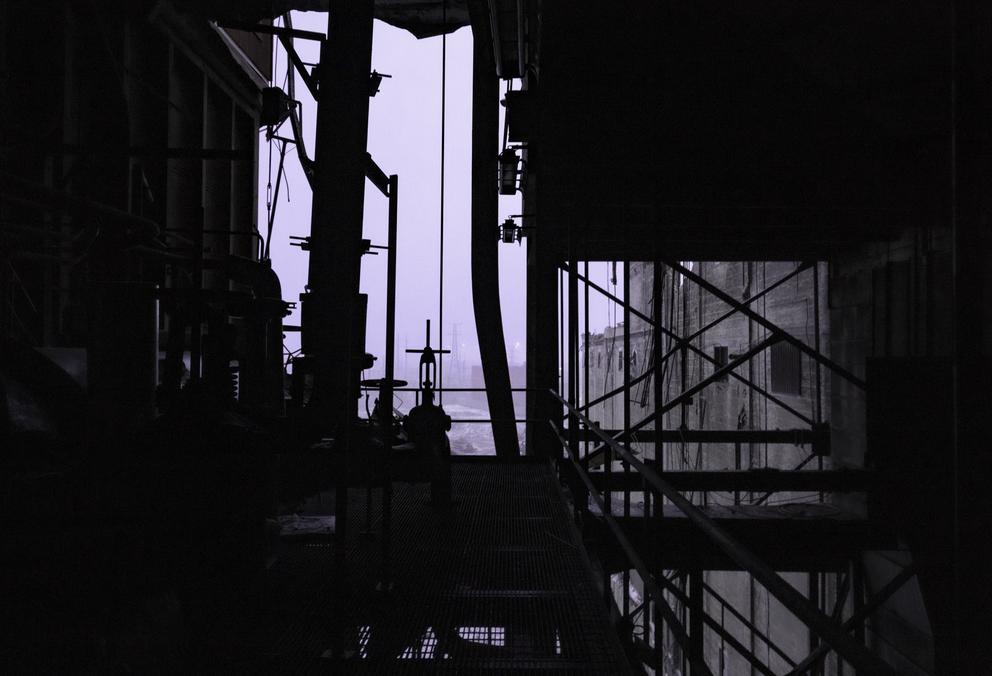 Pipes, valves and scaffolding inside a plant reduced to silhouettes against a pale violet opening